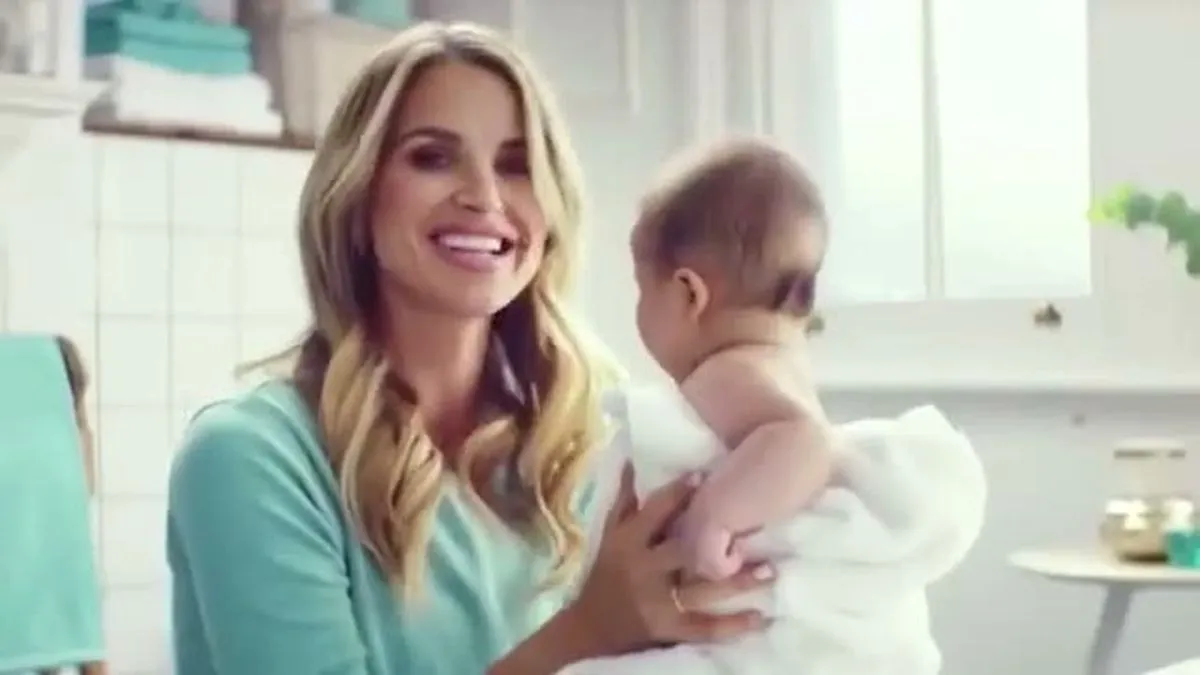 A woman with long blonde hair smiles at the camera while holding a baby wrapped in a white towel.