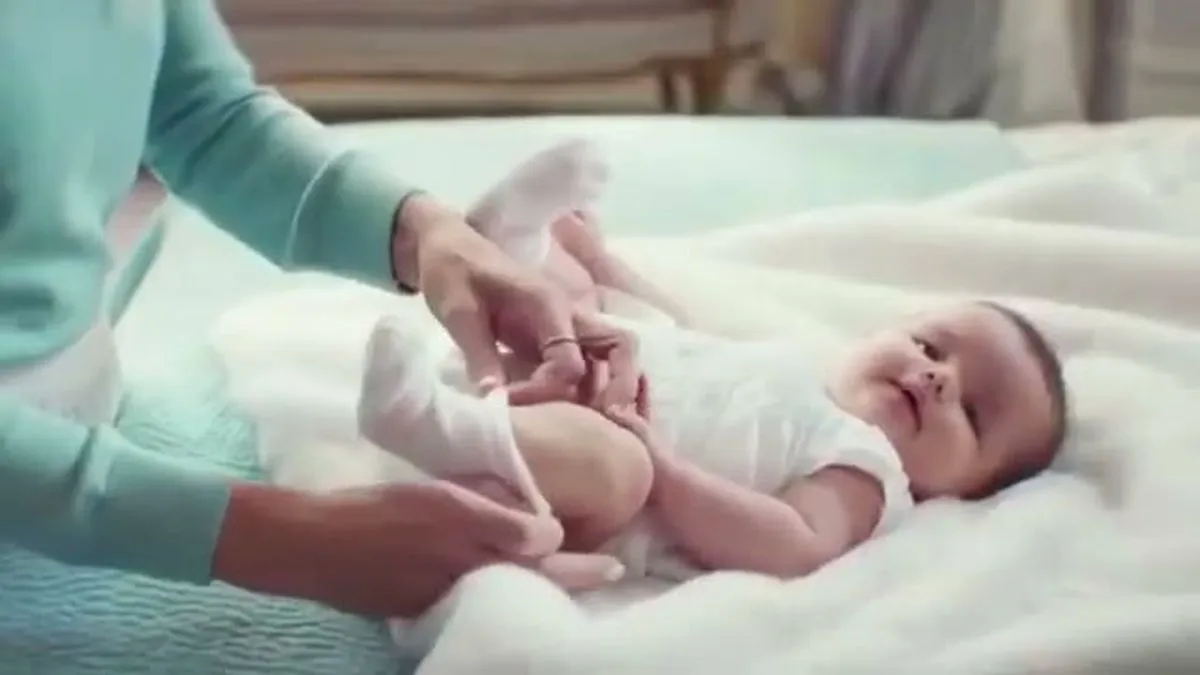 A woman in a teal top and white skirt dresses a baby in a white onesie on a fluffy white blanket.