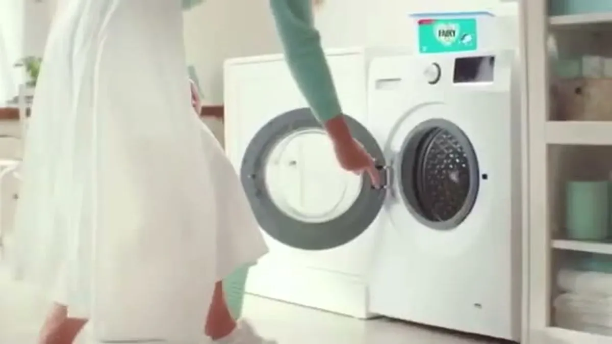 A woman in a teal top and white skirt opens a washing machine door in a bright laundry room.