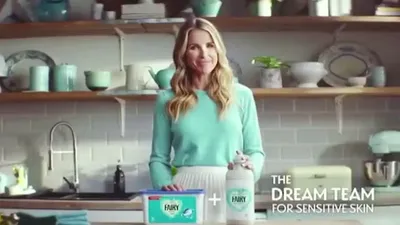 A smiling woman with long blonde hair stands in a kitchen, holding Fairy Non Bio products.