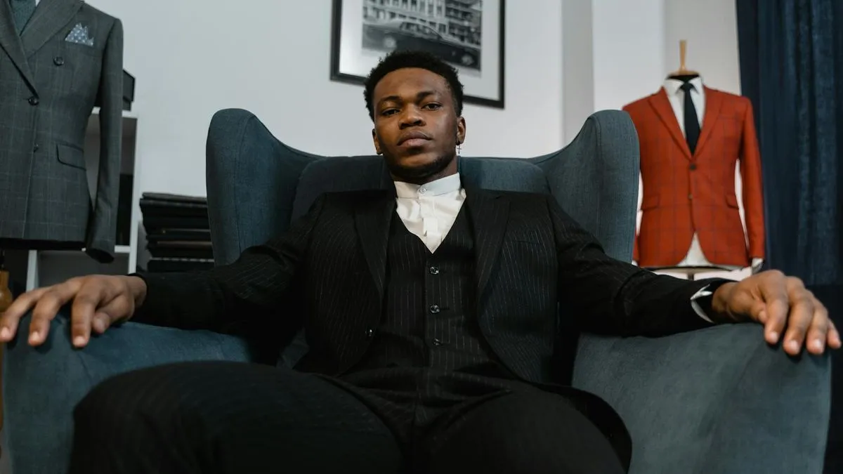 A man in a black suit and white shirt sits confidently in a large armchair.