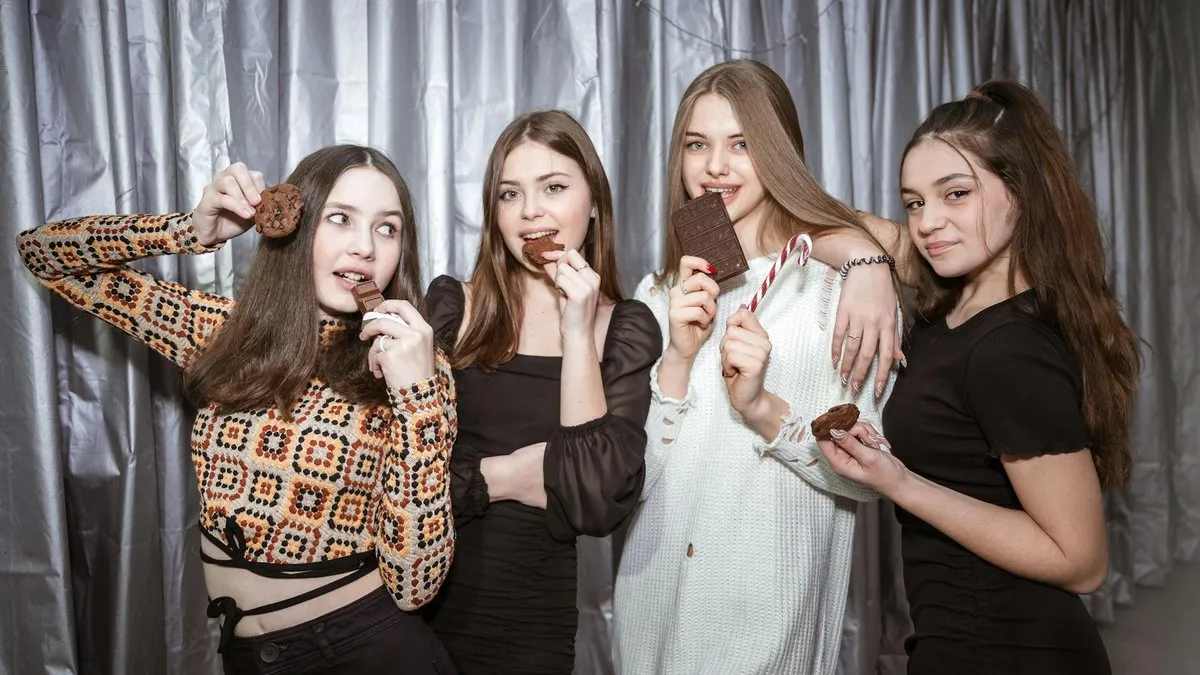 Four young women holding and eating chocolate bars and cookies against a silver background.