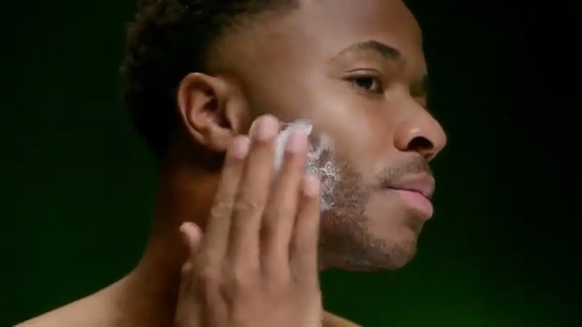 Raheem Sterling applies shaving cream to his face with his hand, looking to the right.