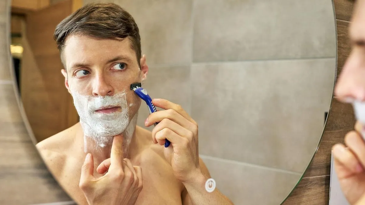 A man with shaving cream on his face shaves with a blue razor in a mirror.
