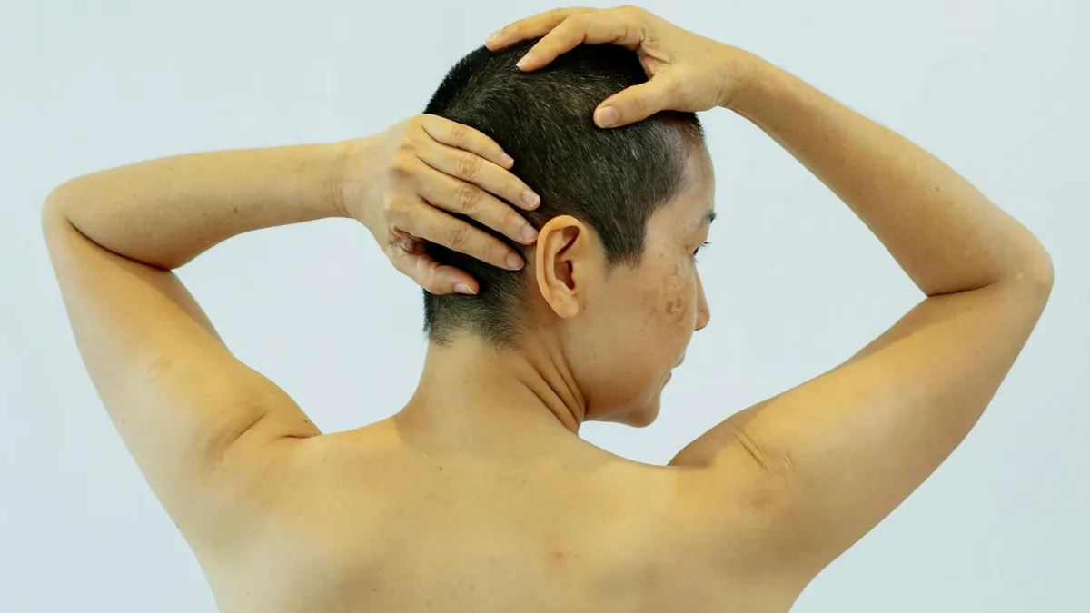 Back view of shirtless woman with short dark hair touching head and looking away against light background