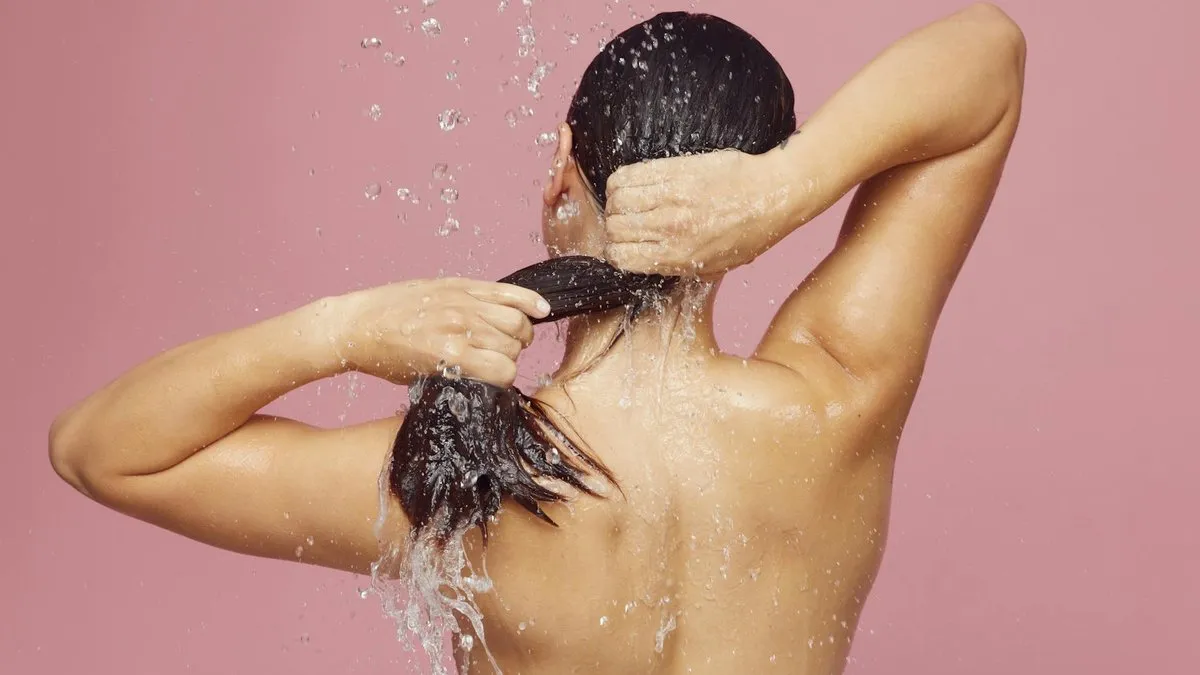 A woman with dark hair washes her hair under a shower against a pink background.