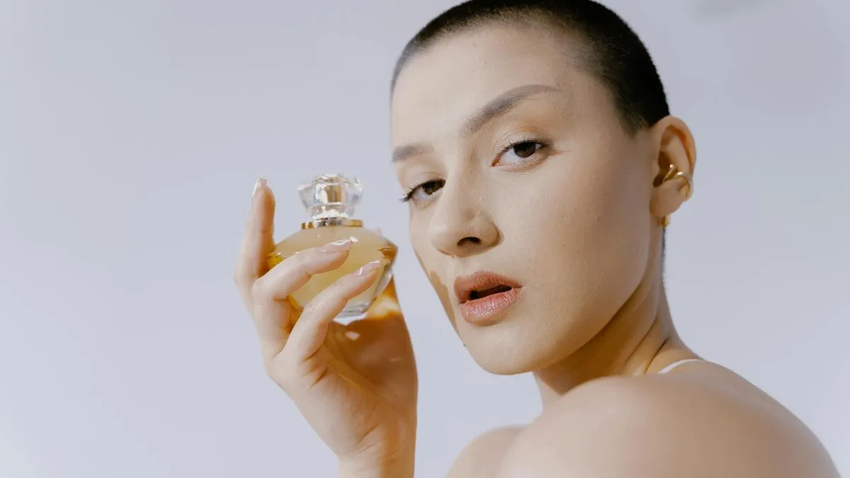 A woman with a buzz cut holding a perfume bottle, looking directly at the camera.