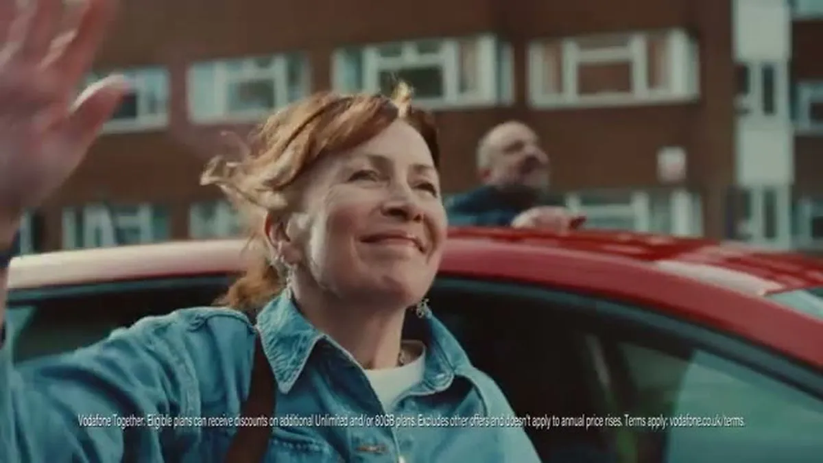 A middle-aged woman with red hair smiles and waves from a red car, with a man waving in the background.