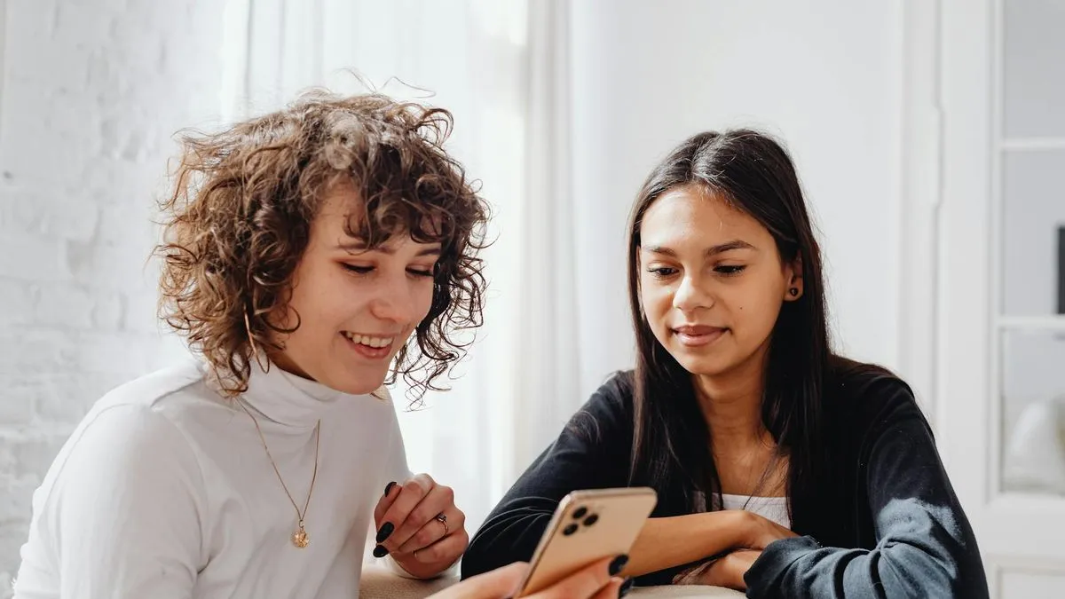 Two young women smiling and looking at a smartphone together in a bright room.