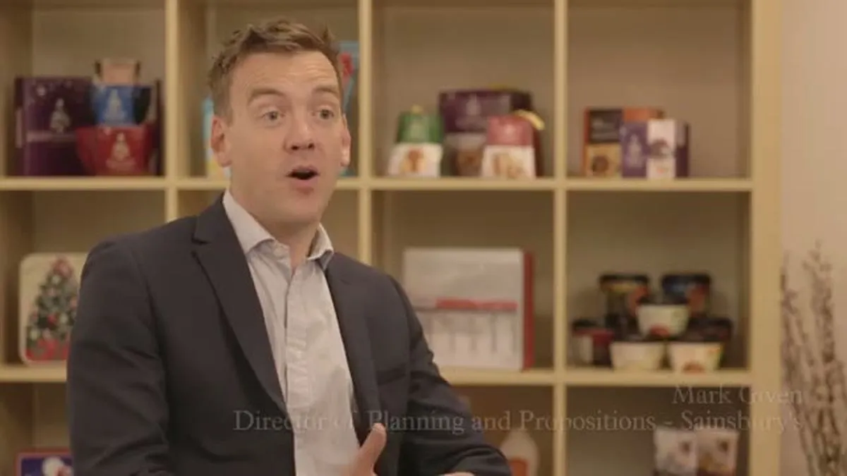 Mark Given, a man in a suit, speaks in front of shelves filled with Sainsbury's products.