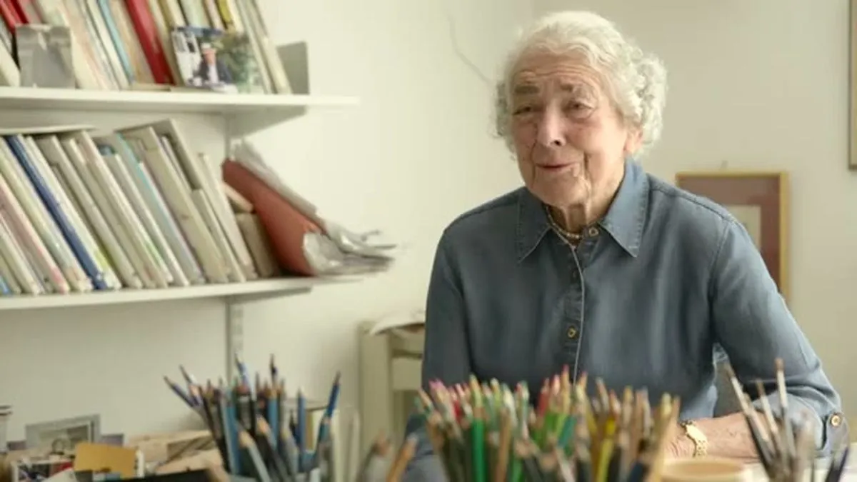 Judith Kerr, an elderly woman with white hair, smiles while sitting at a desk with art supplies.