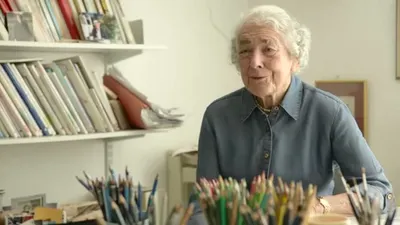 Judith Kerr, an elderly woman with white hair, smiles while sitting at a desk with art supplies.