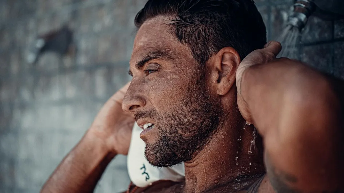 A man with a beard and wet hair is showering, looking up with soap on his face.