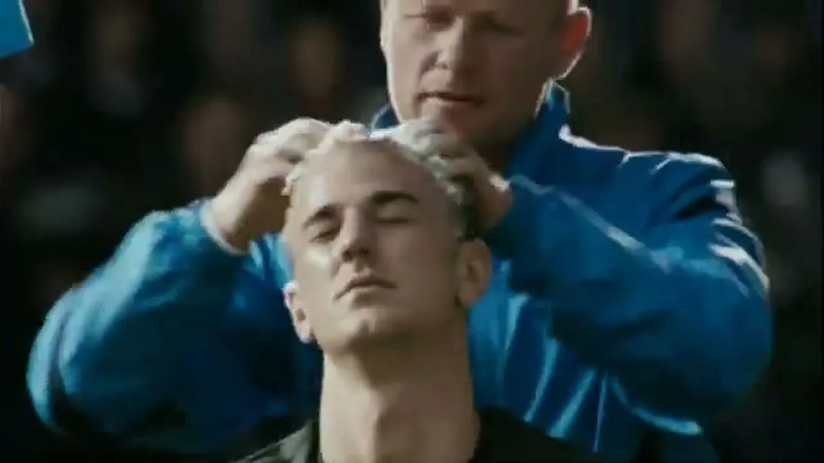 A male footballer with short blonde hair has his hair massaged by a male coach in a blue jacket.