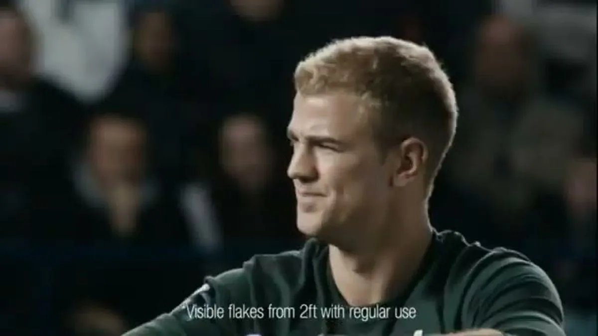 Joe Hart, a male footballer with short blonde hair, looks to the right, smiling slightly, on a football pitch.