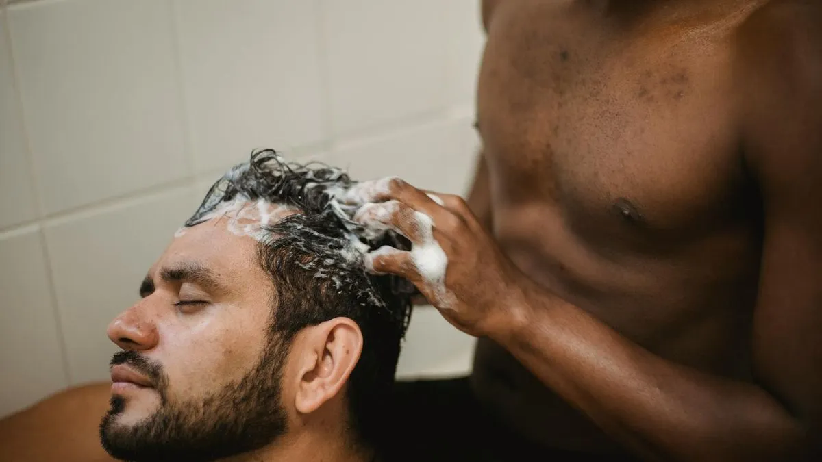 A man with a beard having his hair washed with shampoo by another person.