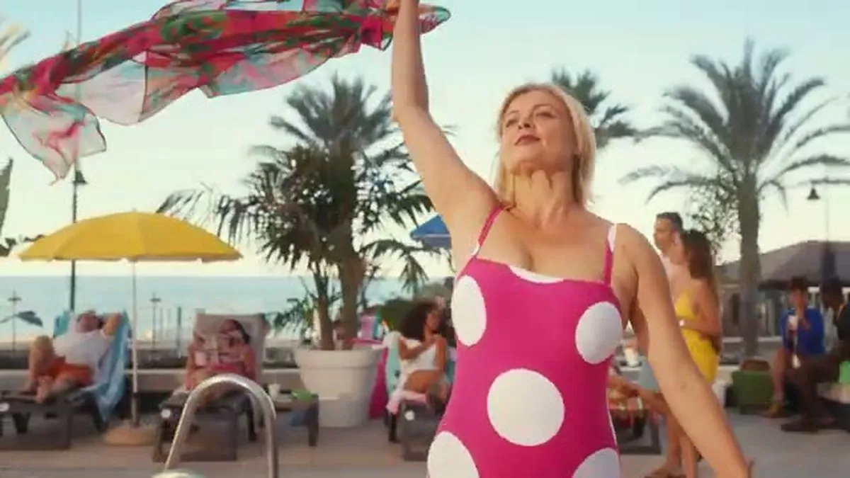 A woman in a pink polka-dot swimsuit stands by a pool, holding a colorful sarong above her head.