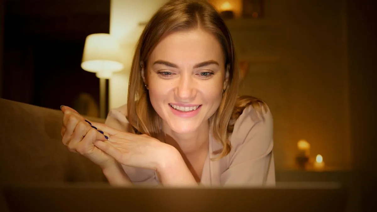 A smiling woman with blonde hair looks down at a screen in a dimly lit room.