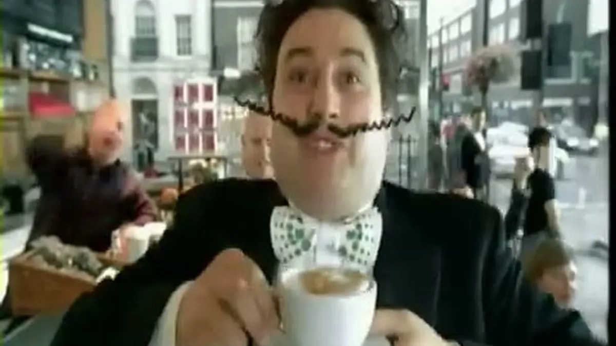 Gio Compario, a man in a tuxedo with a curly mustache, holds a coffee cup and saucer, looking at the camera.