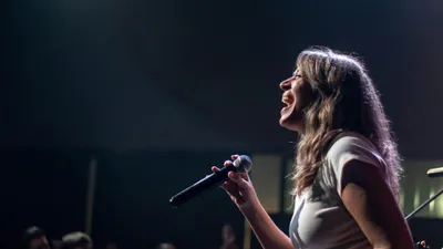 A woman with long brown hair sings into a microphone on a dark stage, illuminated by a spotlight.