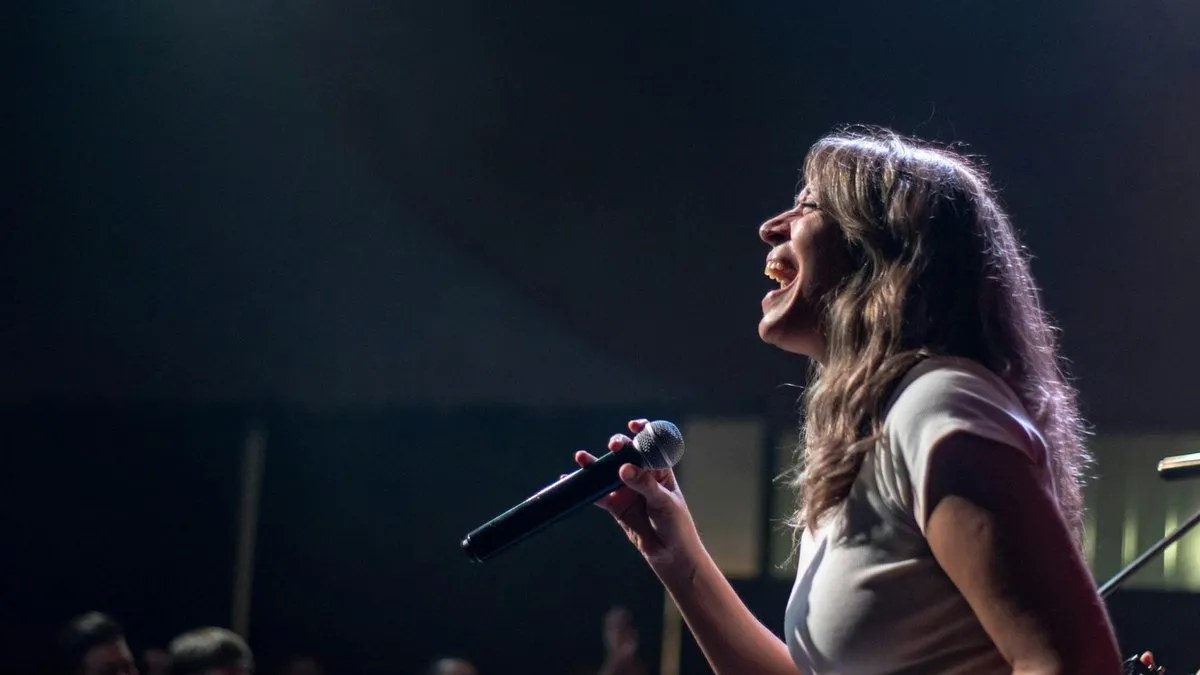 A woman with long brown hair sings into a microphone on a dark stage, illuminated by a spotlight.