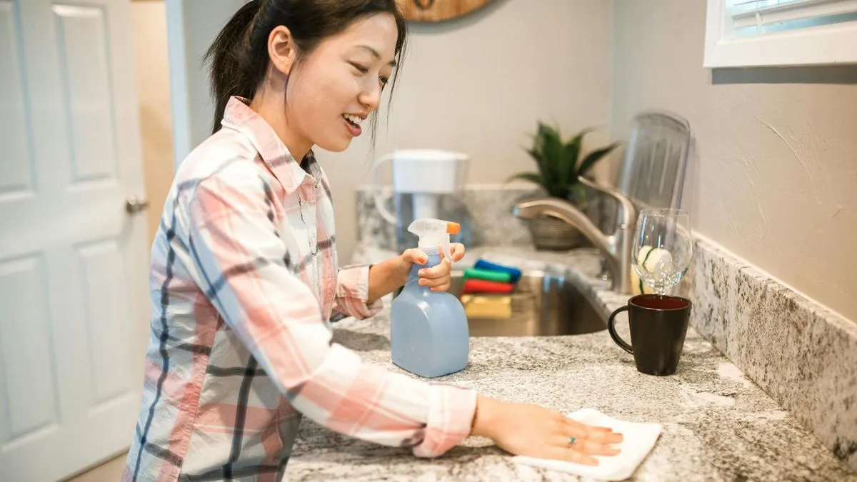 A woman in a plaid shirt smiles while cleaning a kitchen counter with a spray bottle and cloth.