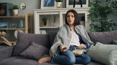A woman wrapped in a striped blanket watches TV on a sofa, holding a remote and popcorn.