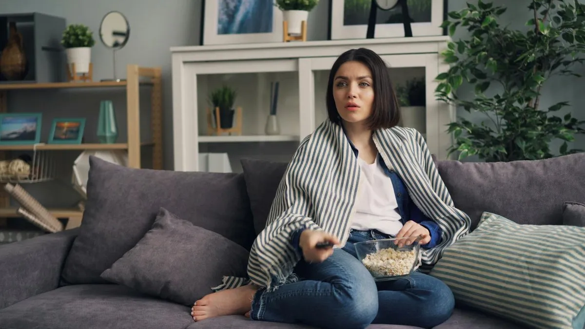 A woman wrapped in a striped blanket watches TV on a sofa, holding a remote and popcorn.