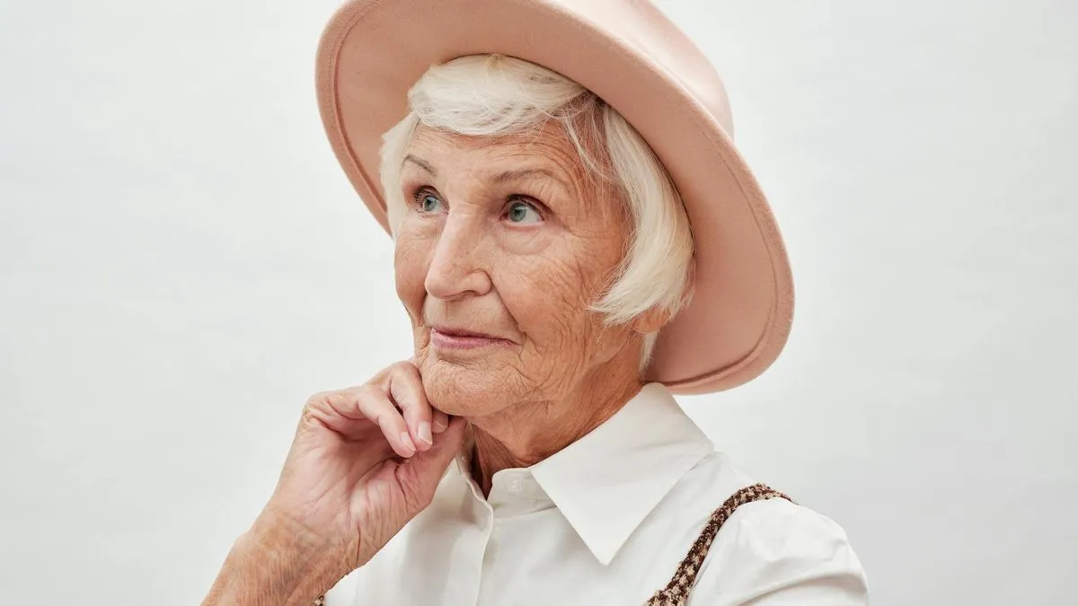 An older woman with white hair and a pink hat looks thoughtfully to the side.