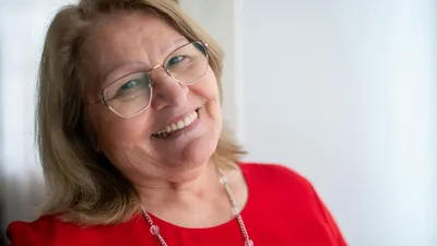 A smiling older woman with blonde hair and glasses wearing a red top and necklace.