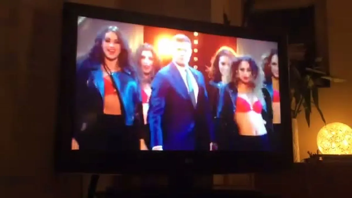 A male presenter in a blue suit stands among several women in black jackets and red bras on a stage.