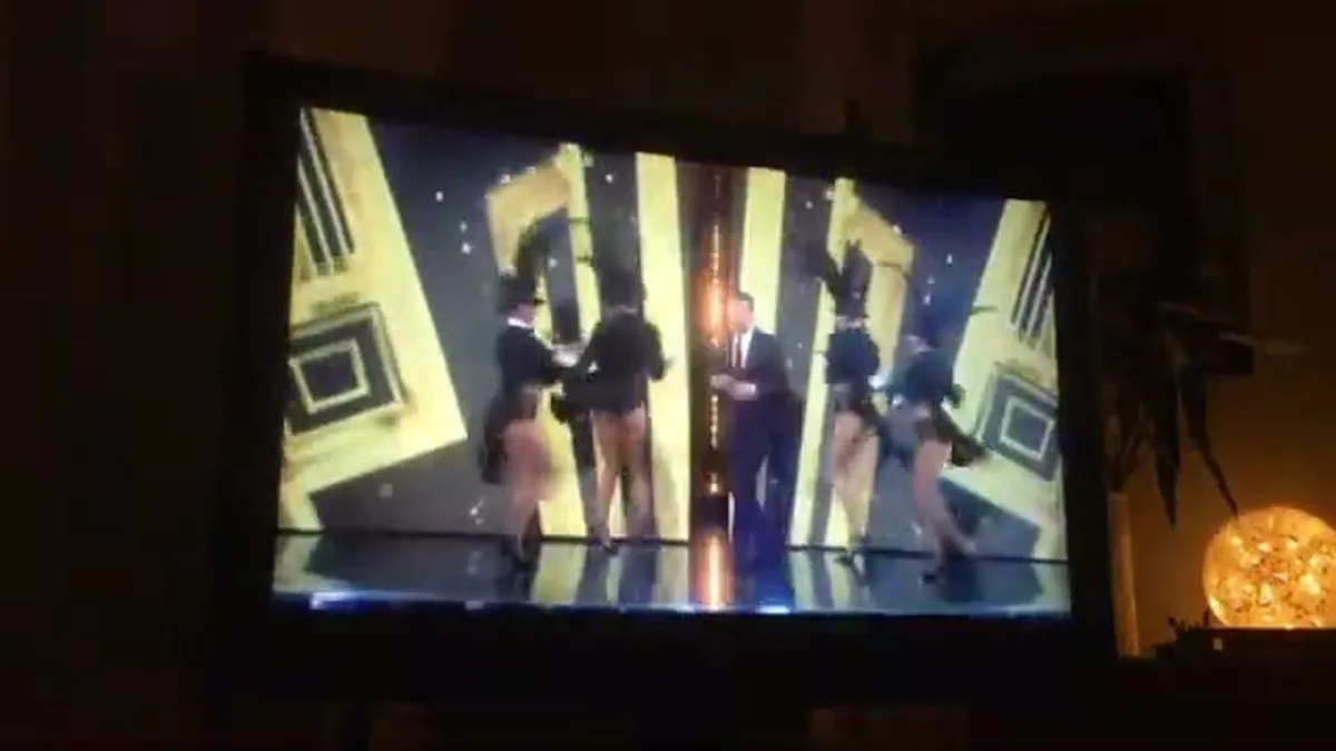 A male presenter in a dark suit stands on a stage with four women in showgirl outfits, against a gold geometric background.