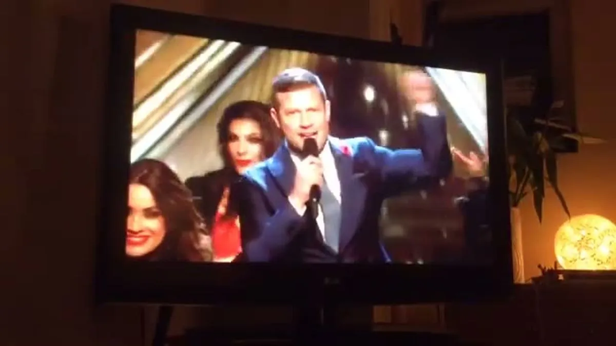 A man in a blue suit sings into a microphone on a TV screen, flanked by two women.