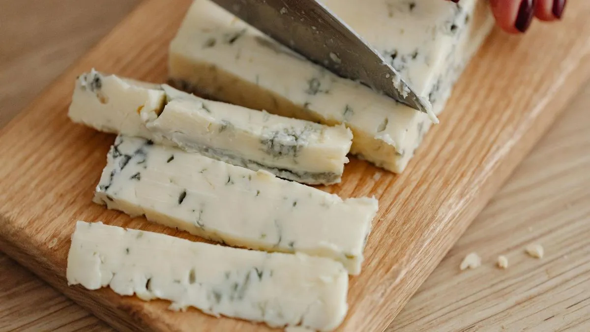 A person's hands slicing a block of blue cheese on a wooden cutting board.