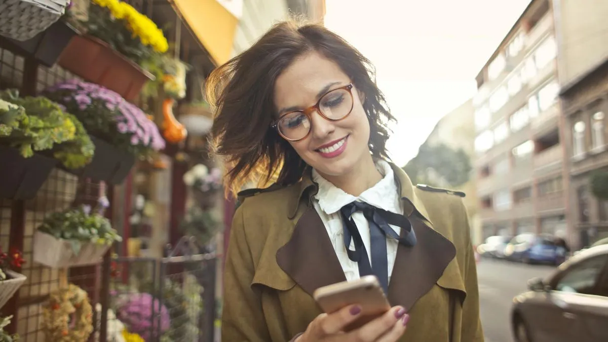 A smiling woman with glasses looks at her phone while walking past a flower shop.
