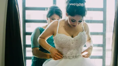 A bride in a white dress with a sparkly belt is being helped by another woman.