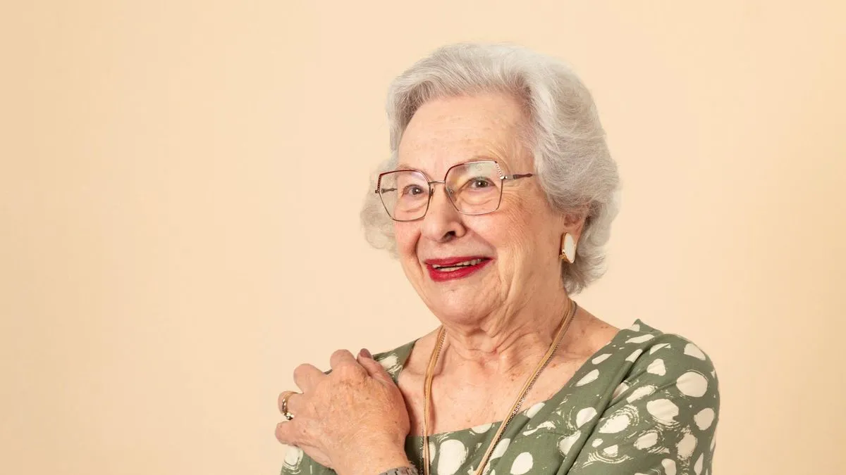 An older woman with white hair and glasses smiles, wearing a green patterned top.