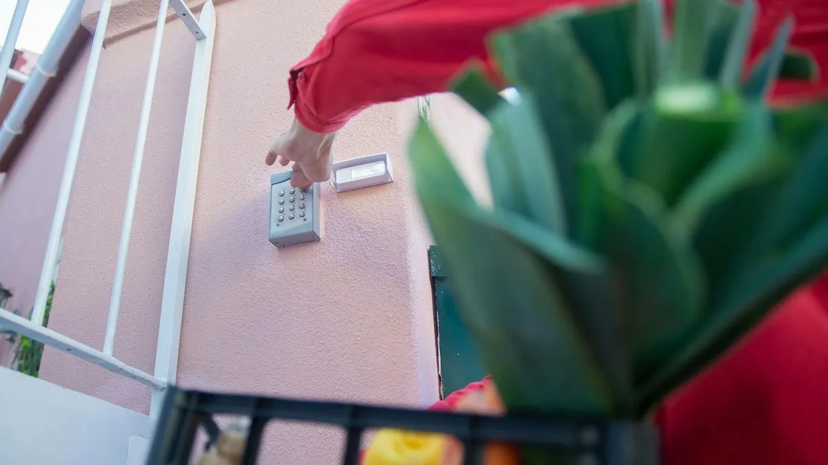 A person in a red jacket presses buttons on an outdoor alarm keypad next to a door.