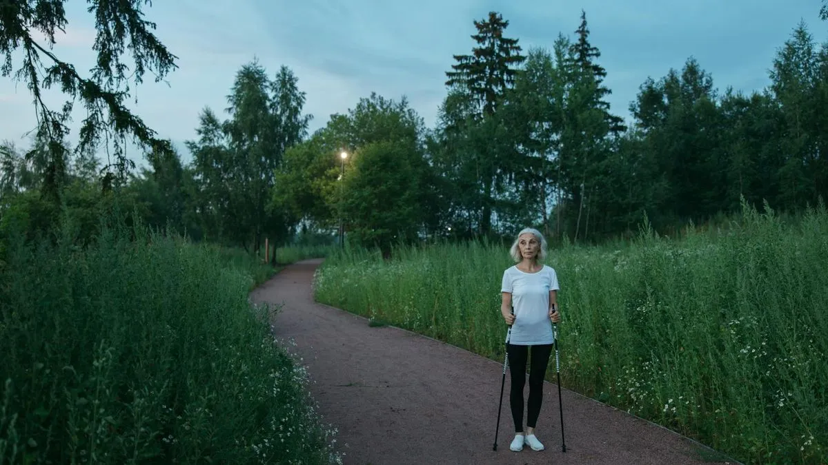 A woman with grey hair in a white t-shirt and black leggings stands on a path with walking poles.