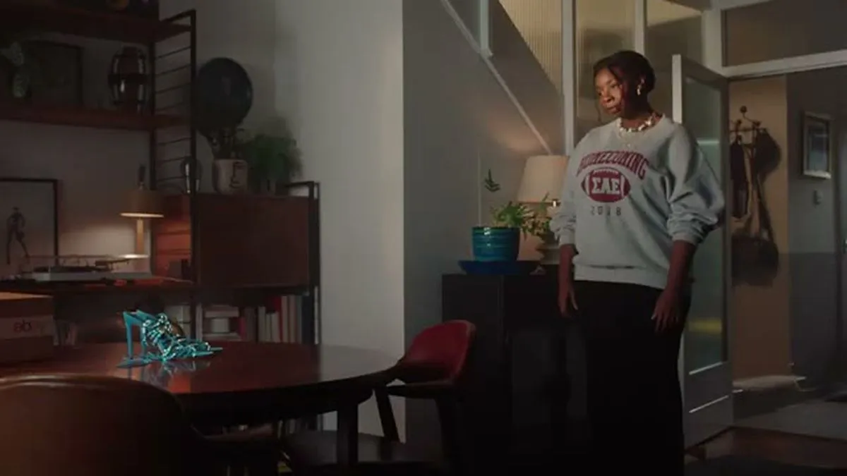 A young Black woman in a sweatshirt stands in a living room with a pair of blue heels on a table.
