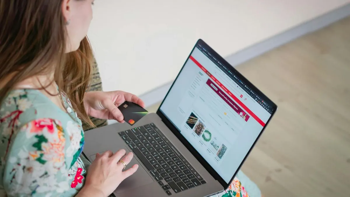 A woman in a floral dress is seen from behind, holding a credit card while using a laptop.