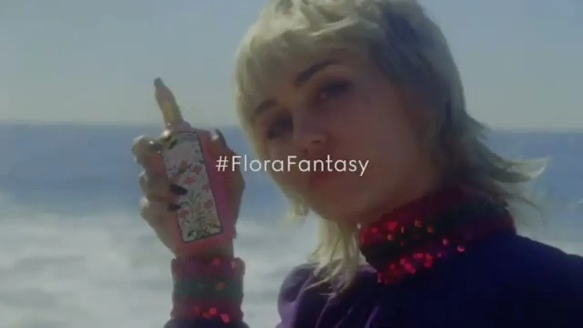 Miley Cyrus holds a Gucci Flora perfume bottle, looking at the camera with the ocean in the background.