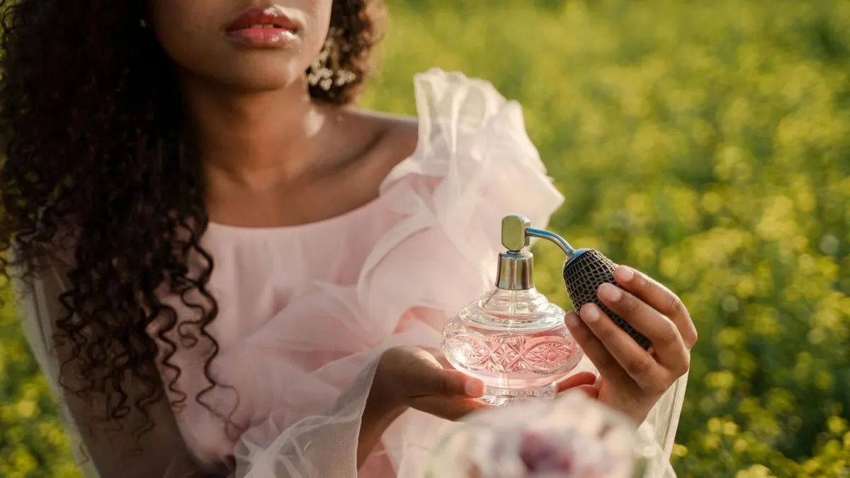 A woman with long curly hair in a pink dress holding a vintage perfume bottle in a field.