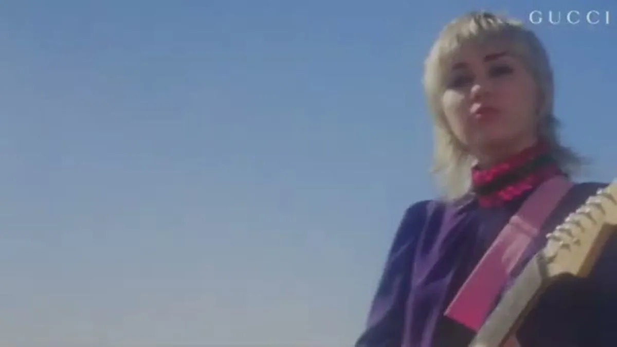 Miley Cyrus, with blonde hair and a mullet, plays a pink electric guitar on a sunny beach.