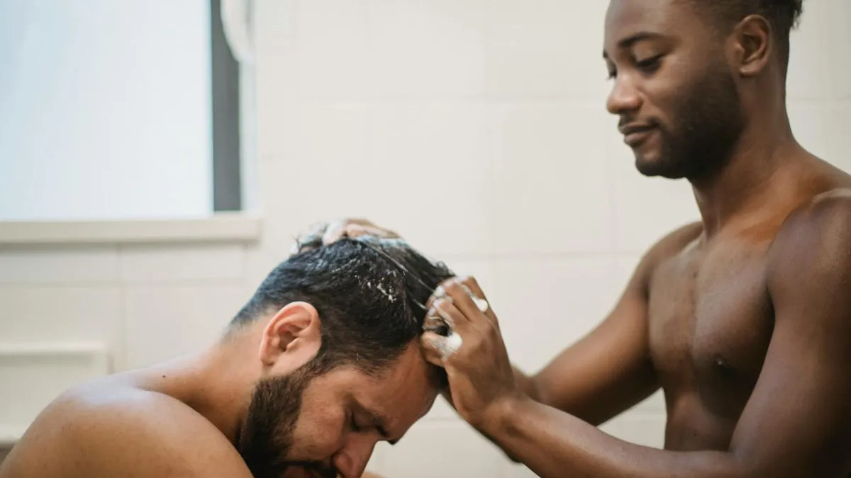 Two men in a bathroom, one washing the other's hair with shampoo and lather.