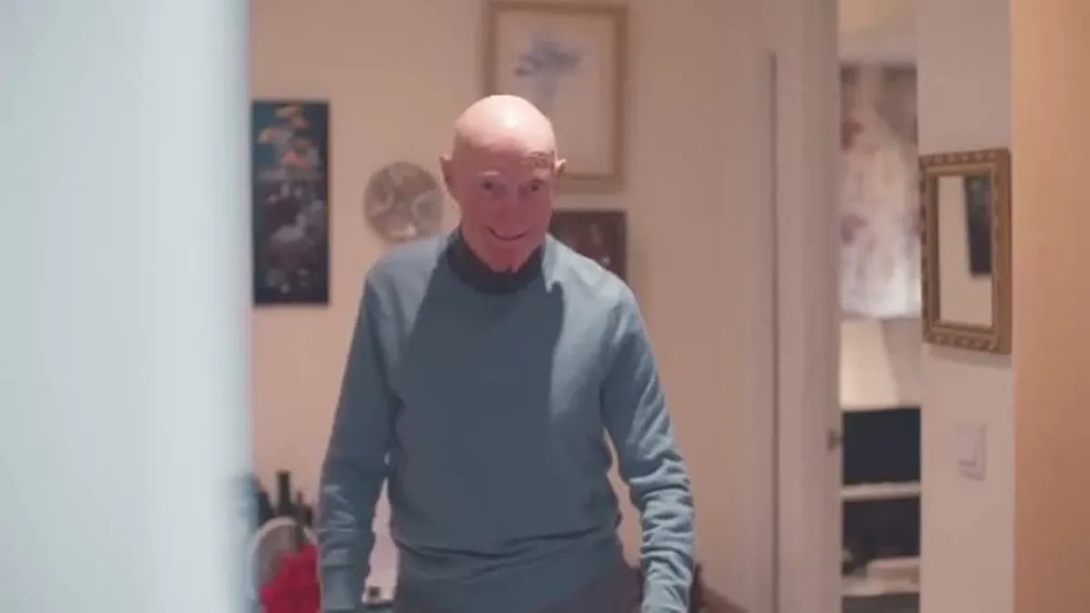 An elderly bald man in a blue sweater walks towards the camera in a hallway.