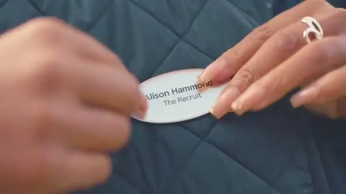 Close-up of hands pinning a name tag that reads 'Alison Hammond The Recruit' onto a quilted vest.