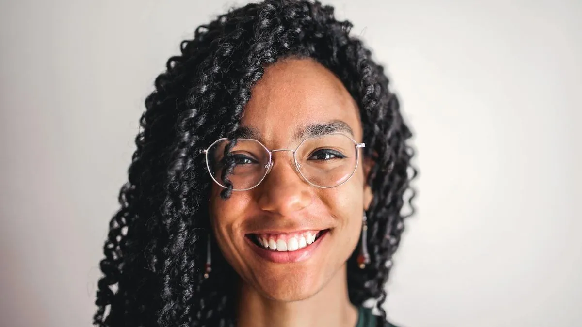 A smiling woman with dark curly hair and glasses looks directly at the camera.