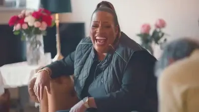 Alison Hammond, a Black woman with a high ponytail, laughs heartily while sitting indoors.