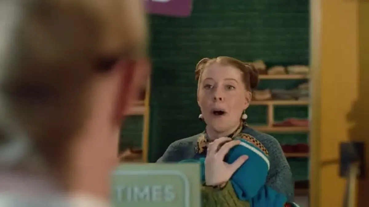 A woman with red hair in pigtails and a green sweater looks surprised, holding a blue knitted item.